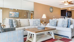 Een woonkamer met een beige muur, een plafondventilator, een flatscreen televisie, een bank met gekleurde kussens, een salontafel en een keukenhoek met een magnetron.