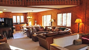 Interior acogedor de una cabaña con una gran mesa de madera para comer, un conjunto de sofás y una cocina con refrigerador y estufa.
