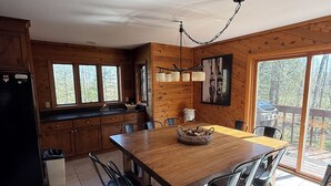 Interior de una cabaña de madera con una mesa de comedor, sillas, un refrigerador y un ventanal grande.