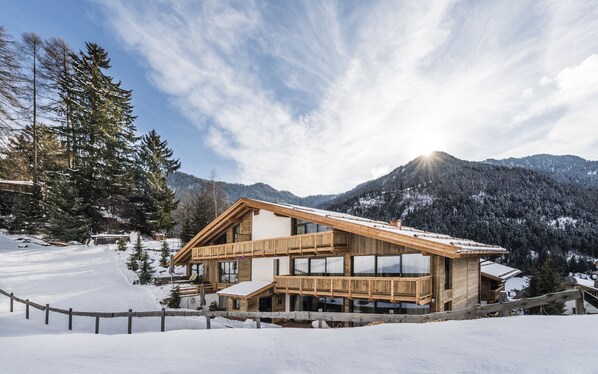 A wooden chalet with a sloped roof, large windows, and a deck, surrounded by snow-covered mountains and trees. - Hotel Montchalet (Ortisei)
