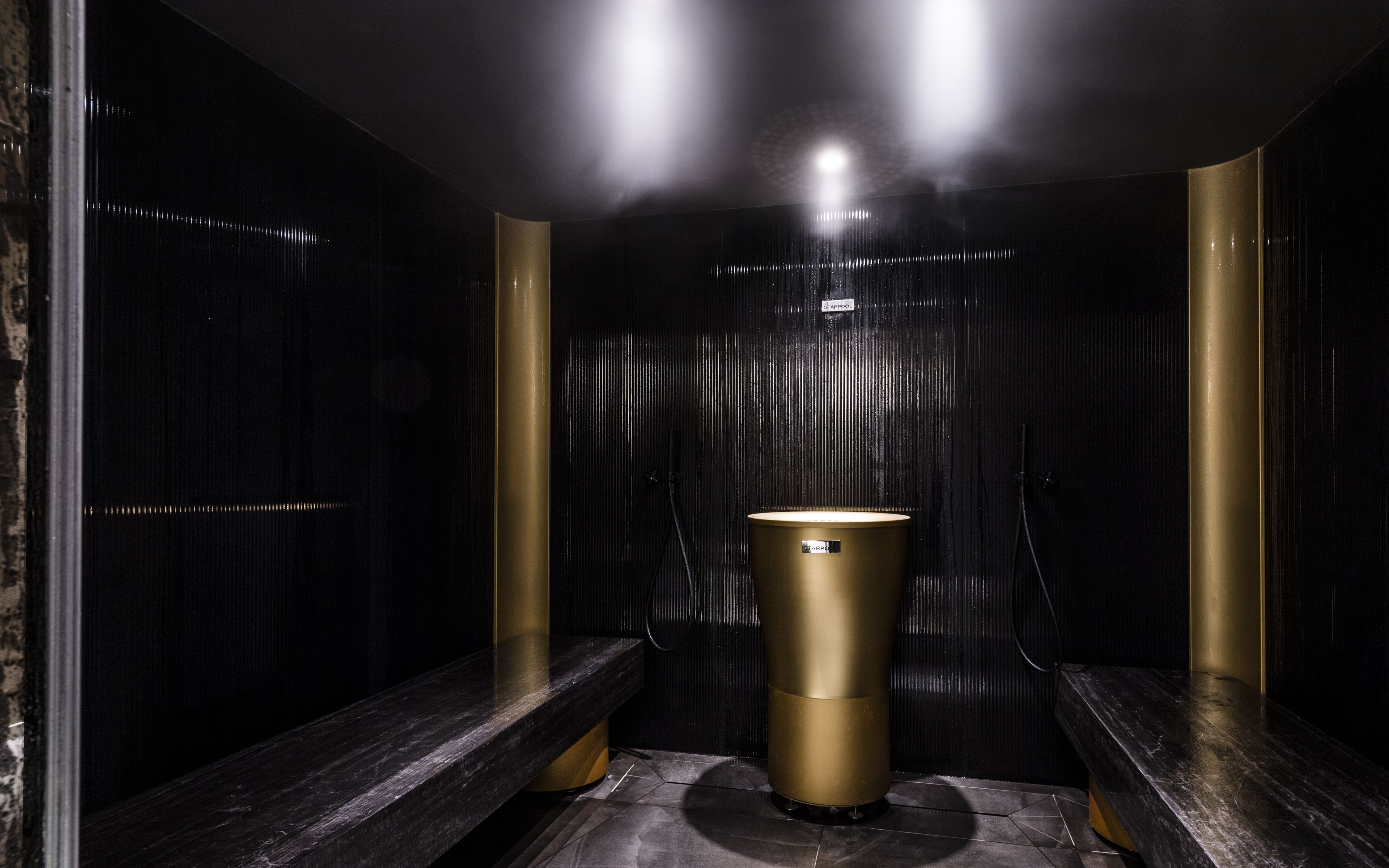 A dimly lit, modern bathroom with a central gold sink, black marble benches, and a textured wall.