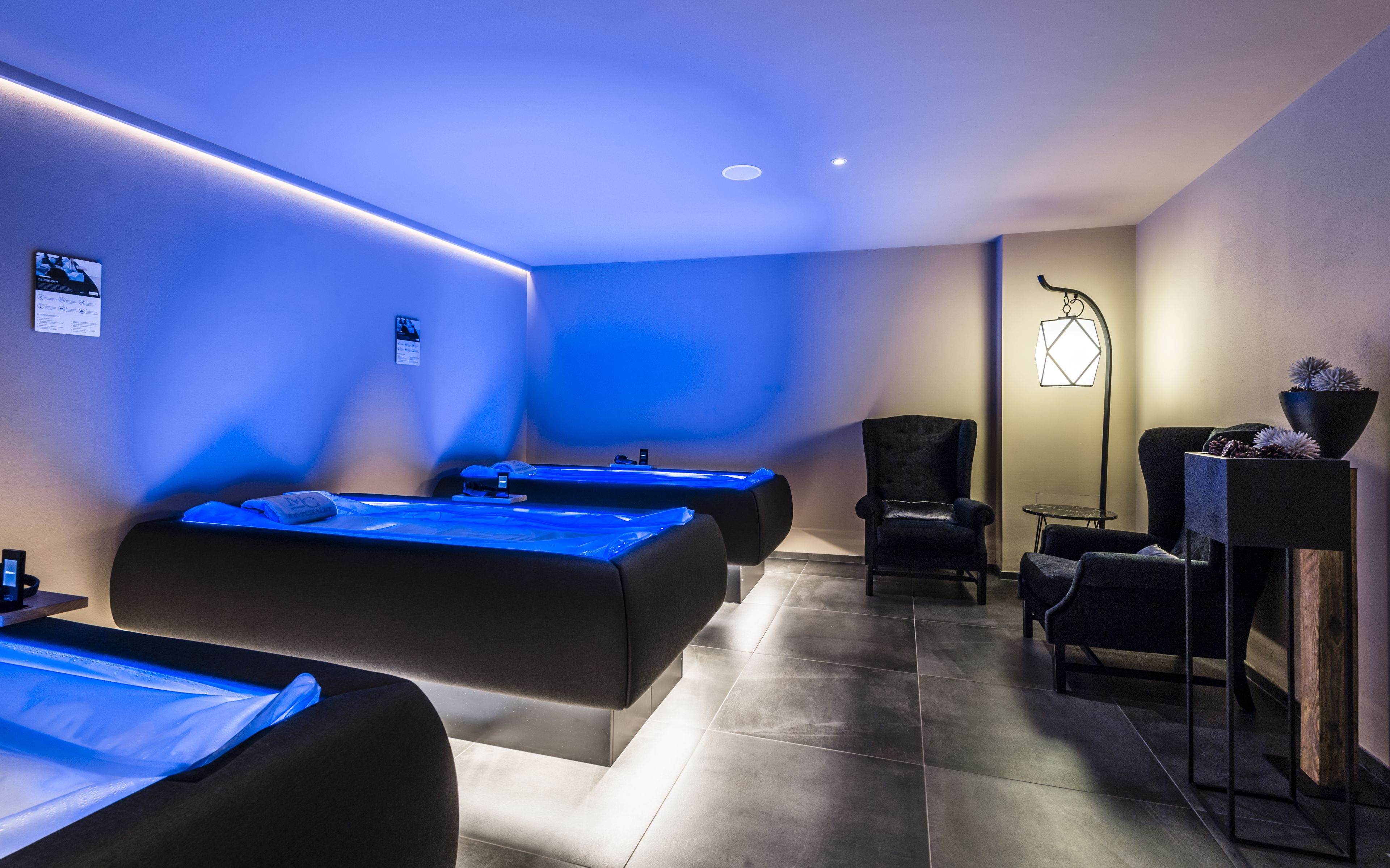 Une salle de spa équipée de deux tables de massage, d’un fauteuil, d’une petite table et d’une lampe décorative.