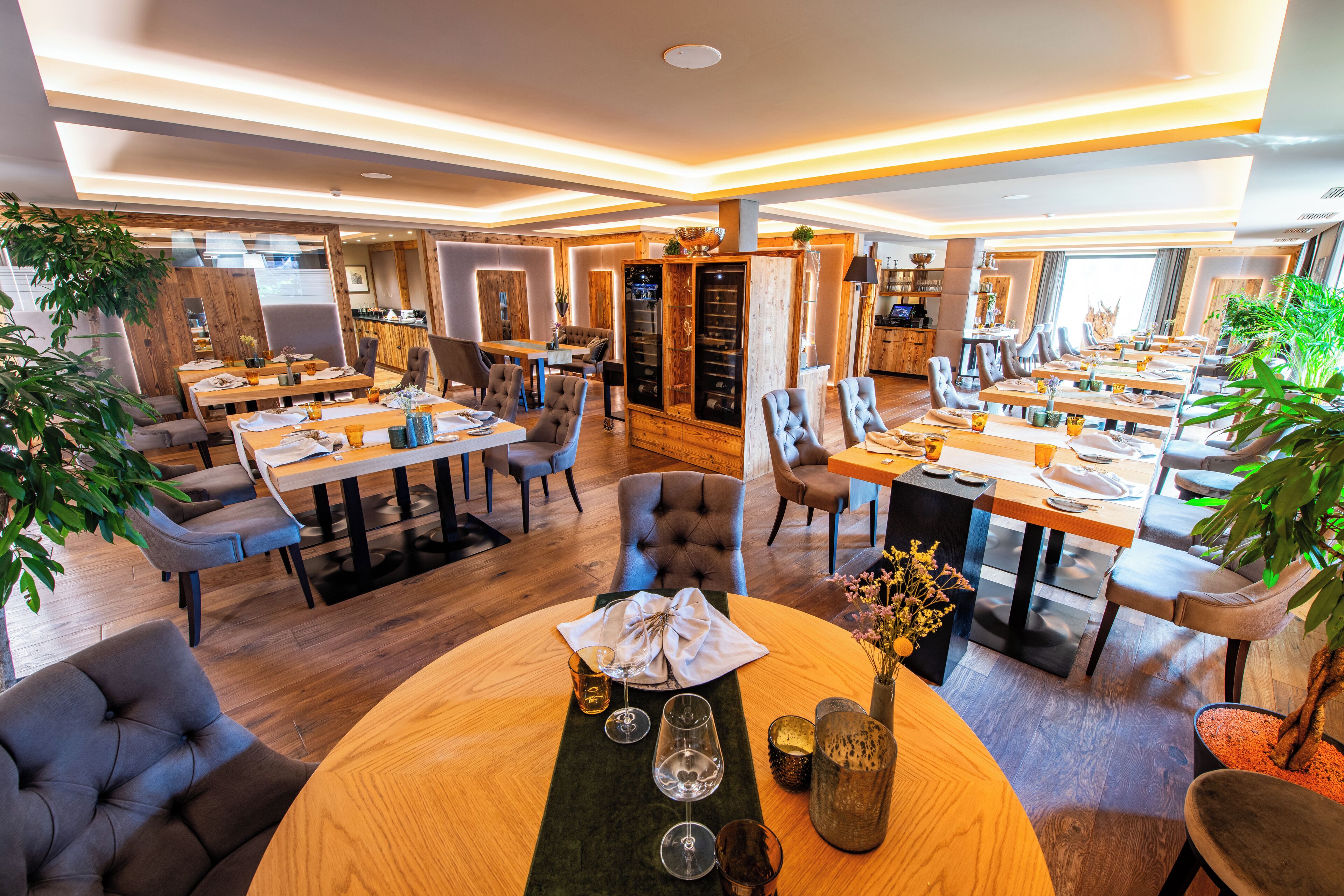 A spacious dining area with multiple tables and chairs, wooden flooring, and potted plants.