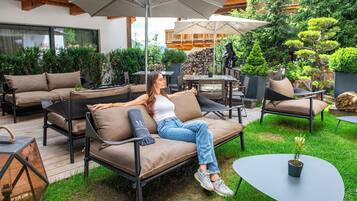 An outdoor seating area with a sofa, chairs, and tables, surrounded by greenery and a stone wall.