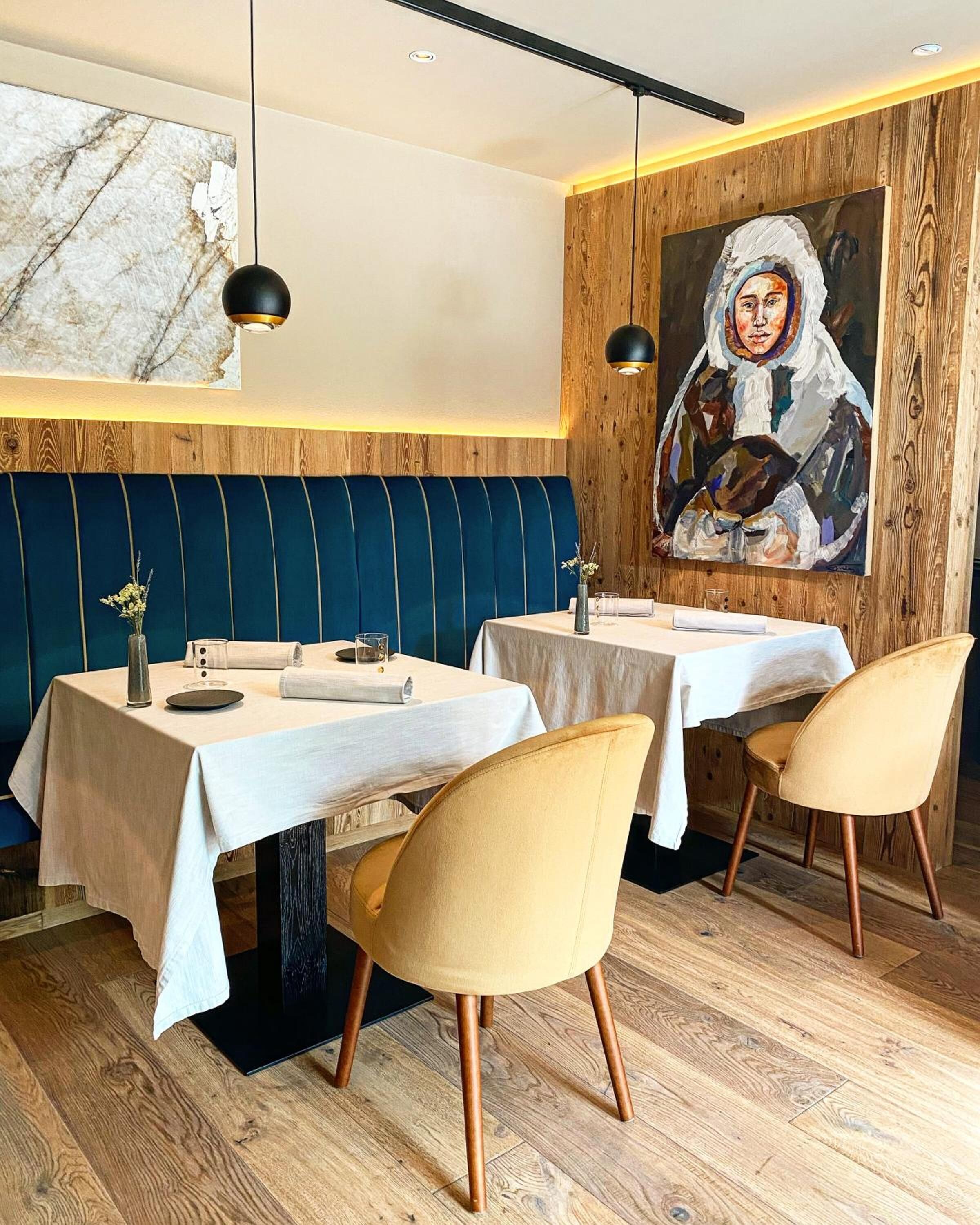 A restaurant with two tables, blue upholstered booths, wooden flooring, and a large painting on the wall.