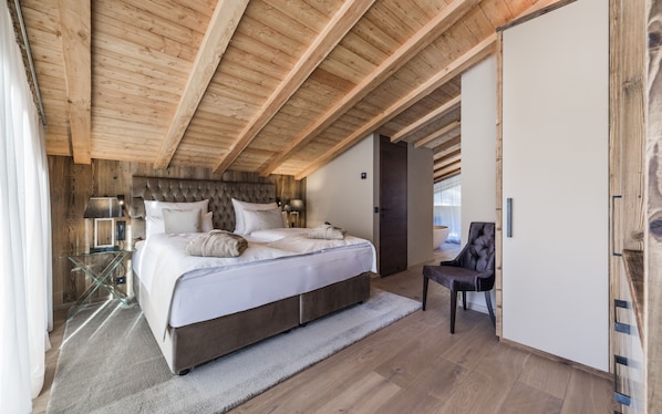 A bedroom with a large bed, wooden ceiling beams, a glass table, and a chair. - Hotel Montchalet (Ortisei)