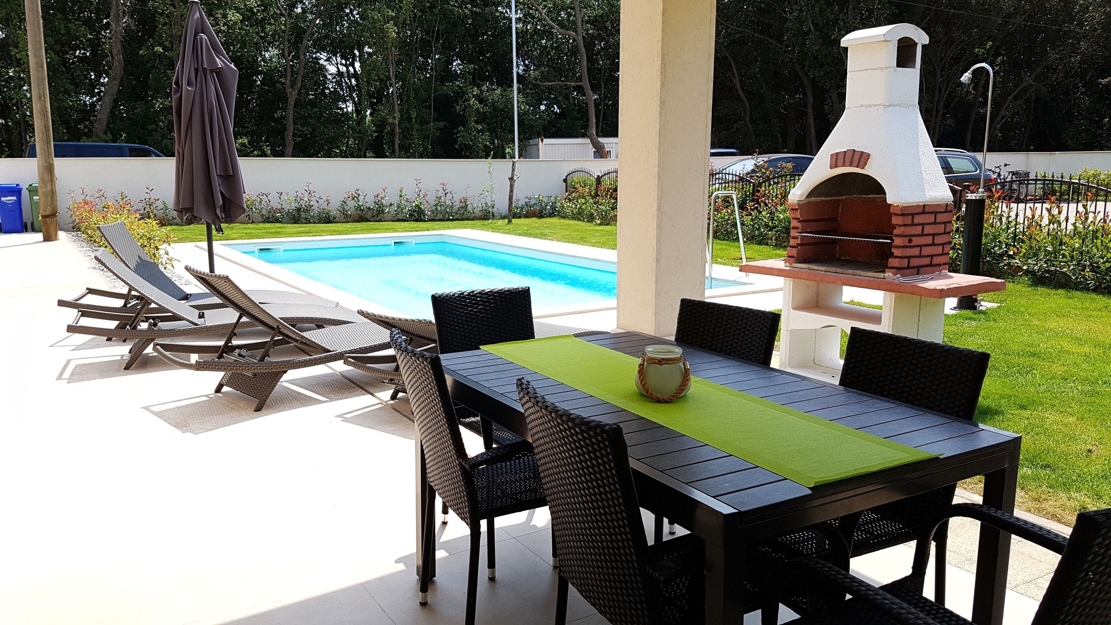 An outdoor patio with a pool, lounge chairs, a table with chairs, and a brick oven.