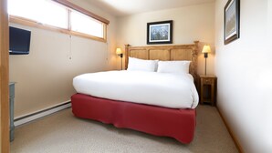 Una cama bien hecha con falda roja, dos lámparas de noche, un cuadro en la pared y un televisor también en la pared.