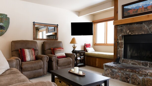 Sala de estar con dos sillones de cuero, una mesa de centro con velas, una chimenea de piedra, un televisor montado y una ventana con vista a la montaña.