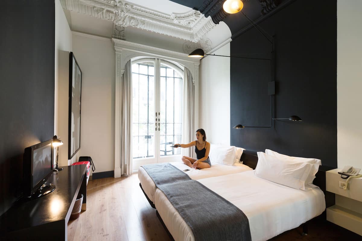 A modern hotel room with a large bed, a desk with a lamp, a TV, and a view of the outdoors through a large window.