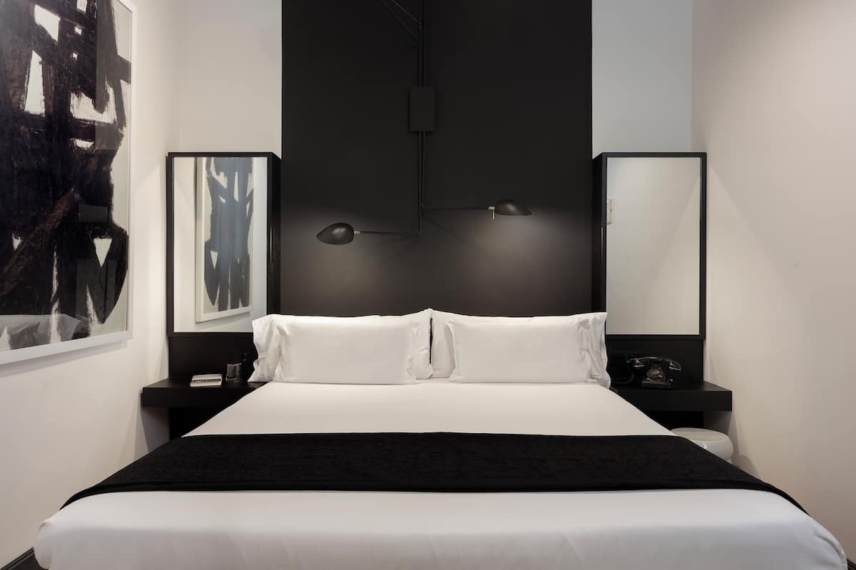 A modern hotel room with a large bed, two bedside tables, a nightstand, a black lamp, and a large abstract painting on the wall.