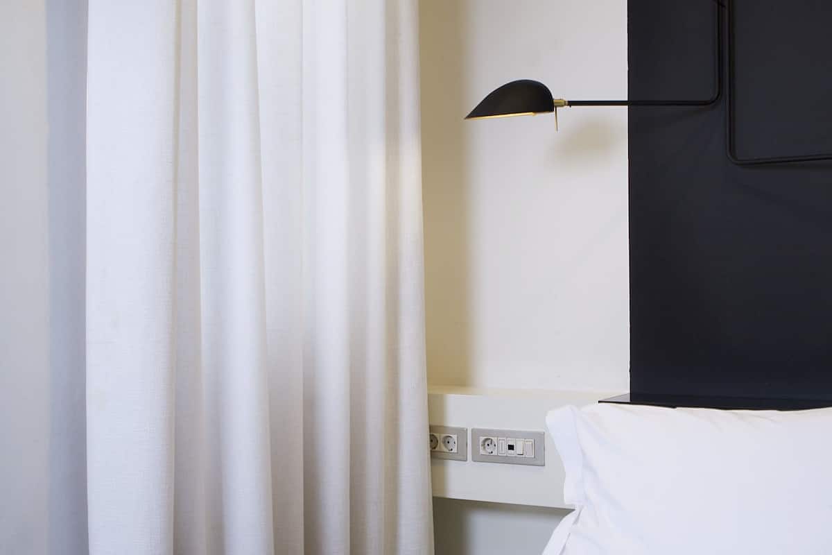 A hotel room with a bed, white curtains, a black wall-mounted lamp, and a white headboard.
