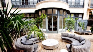 Un espace extérieur circulaire aménagé avec des meubles en osier et une table ronde centrale, entouré de plantes en pot et, au fond, d’un bâtiment doté de balcons.