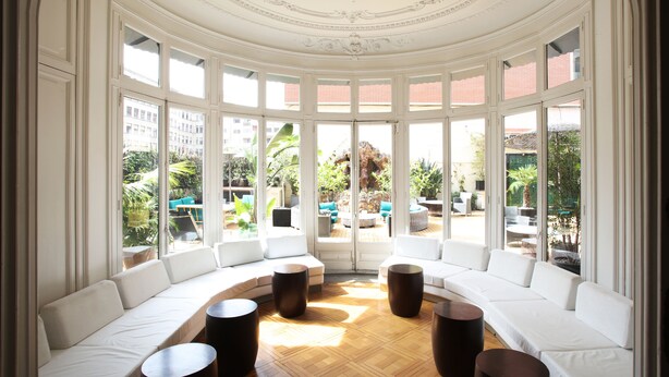 Une pièce baignée de lumière, avec un plafond blanc aux lignes courbes, de grandes fenêtres et un canapé blanc agrémenté de petites tables rondes.
