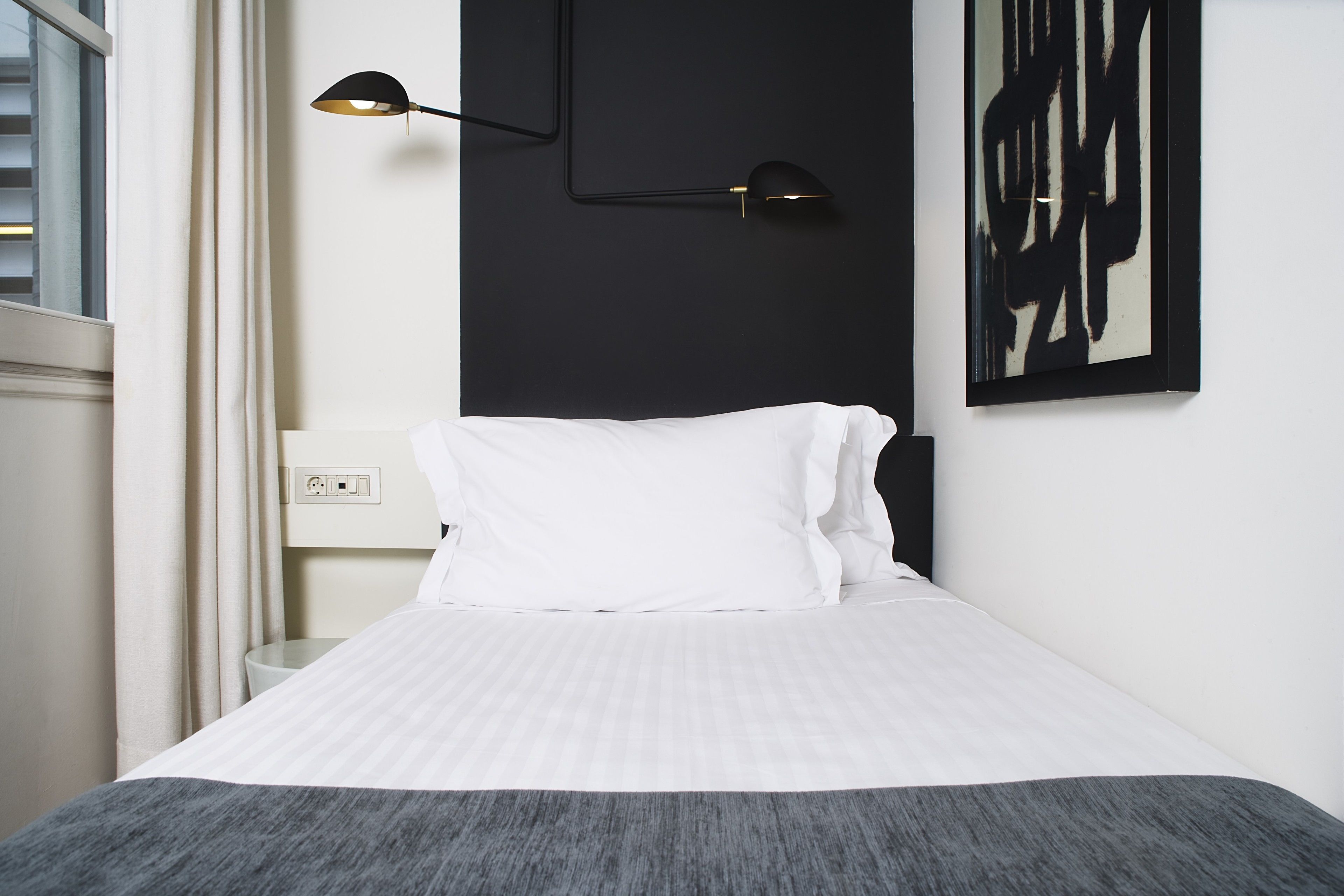 Una cama bien tendida con ropa de cama blanca y un plaid gris, una obra de arte negra en la pared y una lámpara negra sobre la mesita de noche.