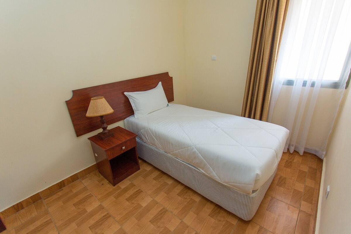 Una cama individual con cabecera de madera, una mesita de noche pequeña con lámpara y una pared lisa.