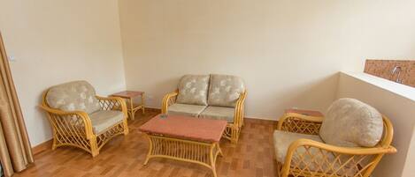 A room with a wooden parquet floor, two wicker sofas with cushions, a small wooden table, and a wall-mounted shelf.