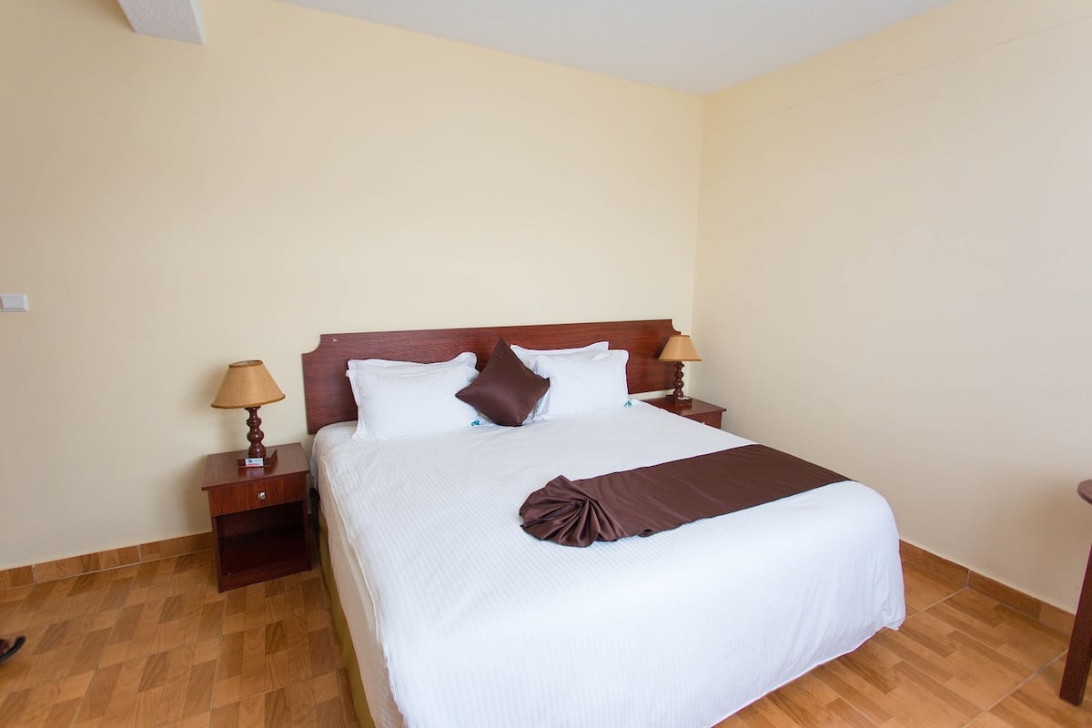 Una cama bien hecha con una manta marrón, dos lámparas de noche y una mesita de madera.