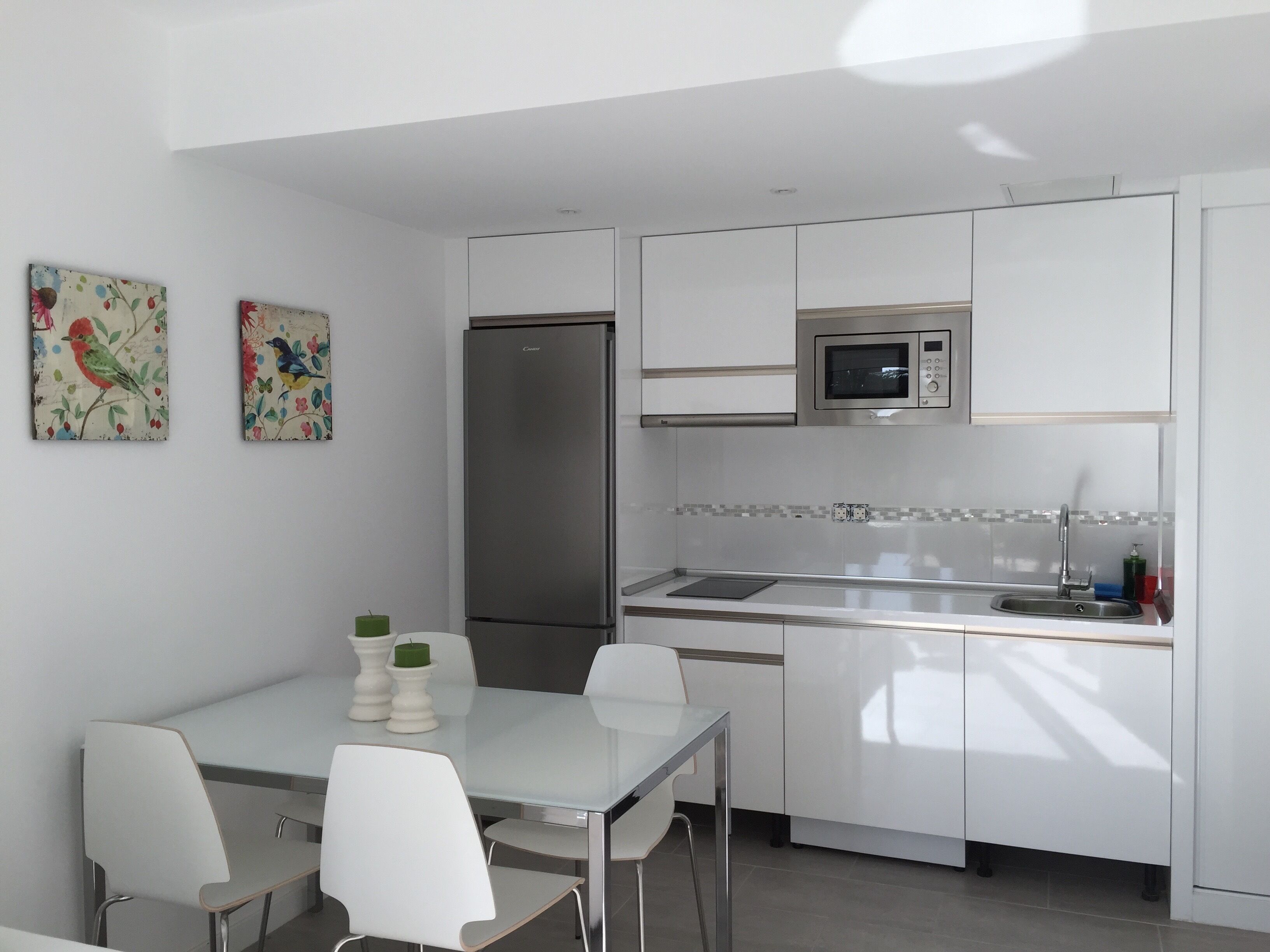 A modern kitchen with white cabinets, a stainless steel refrigerator, a microwave, and a sink. There are two chairs around a table with a vase and candles.