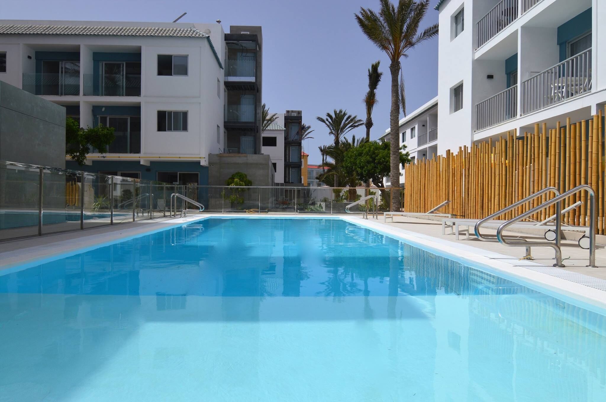 A swimming pool with a wooden fence and metal railings, surrounded by modern buildings with balconies and palm trees.