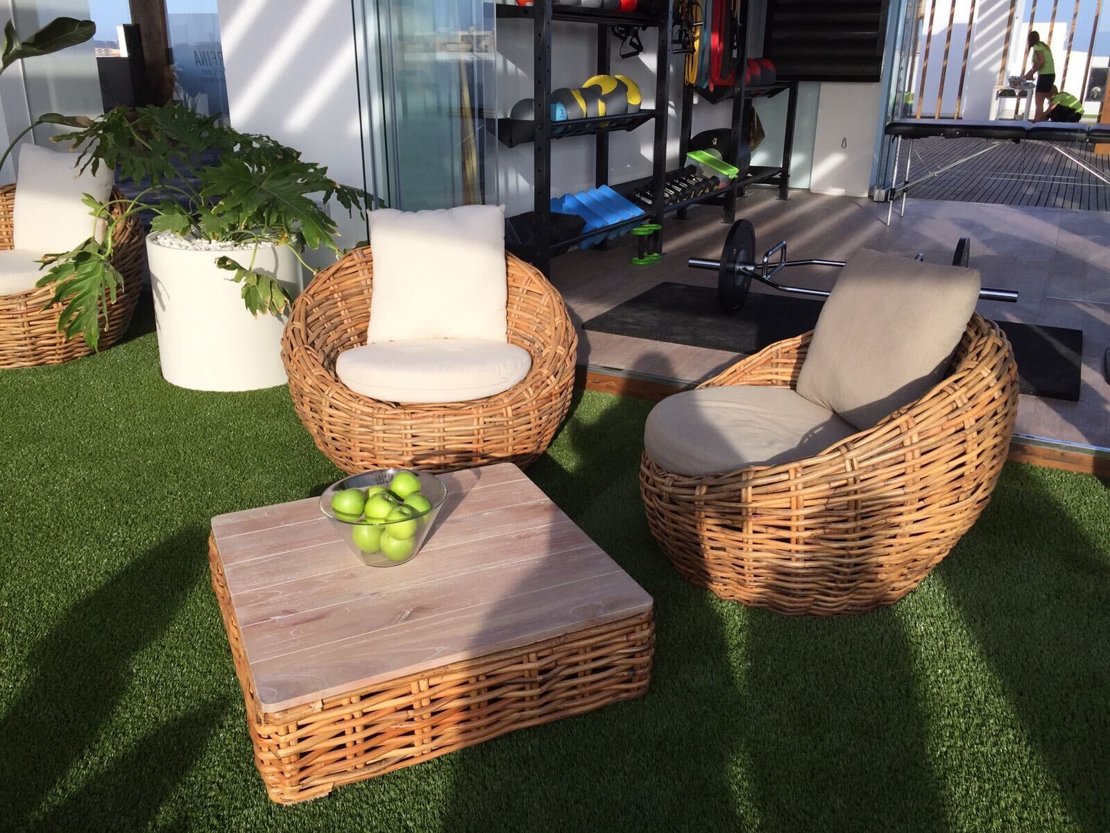 A sunlit outdoor seating area with wicker furniture, a small table with apples, and a gym in the background.