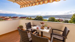 A balcony with wicker chairs, a table with plates of food, and a view of a lake and houses. - Apartment with sea view and quiet Mediterranean ambience (Dobrinj, Krk)
