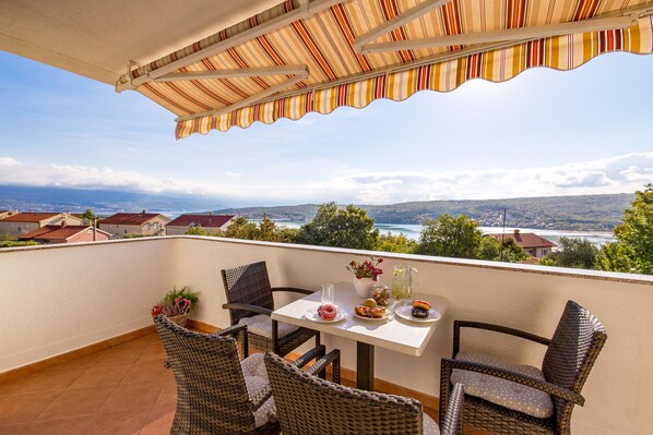 A balcony with wicker chairs, a table with plates of food, and a view of a lake and houses.