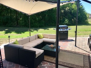 Patio esterno con divano componibile, barbecue e pergola decorata con luci a stringa.