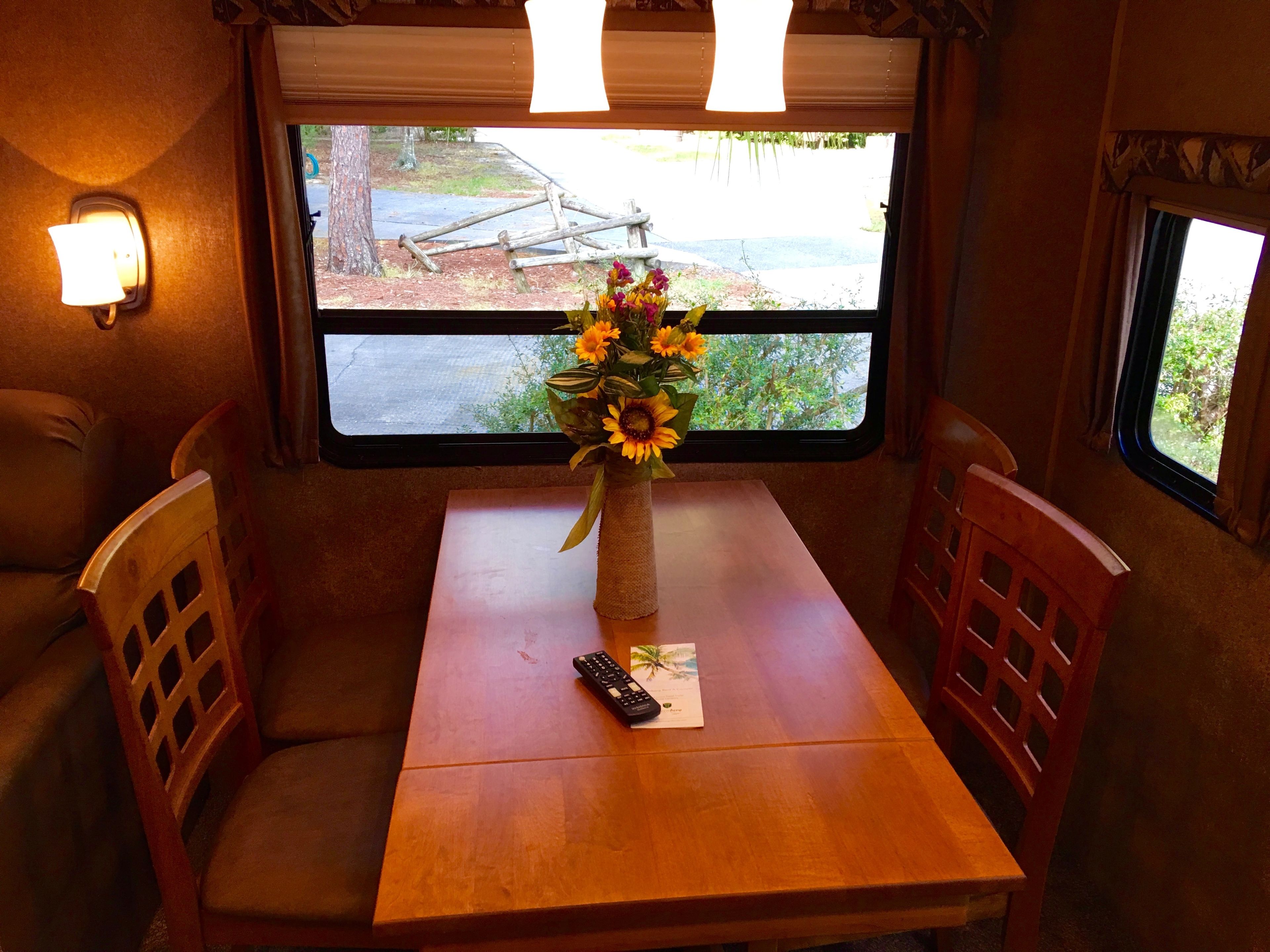 A table with a vase of flowers, a remote control, and a menu on a wooden table in a room with a window.