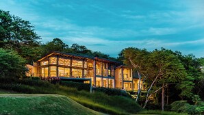 Ein modernes, zweigeschossiges Haus mit großen Fenstern und einer überdachten Terrasse, umgeben von üppiger Vegetation und einem Golfplatz.