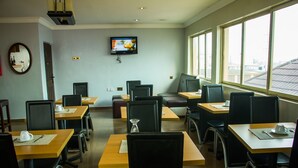 Un comedor con mesas y sillas, un televisor montado en la pared y ventanas amplias.