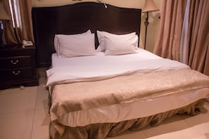 A neatly made bed with white linens and a beige bedspread, a dark wooden headboard, a bedside table with a lamp, and a telephone. - Tivoli Residence & Bar (Lagos)