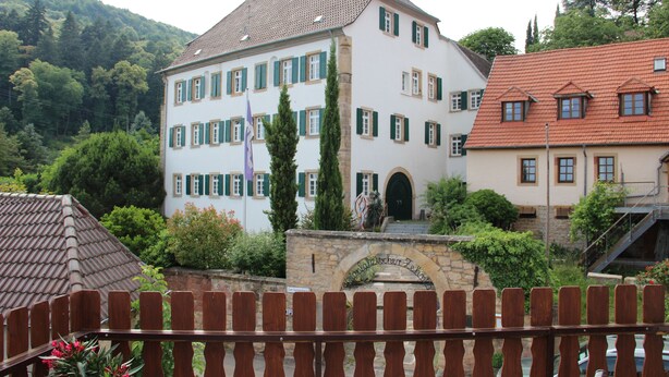 Un immeuble à plusieurs étages, doté d’une entrée en arc de pierre, entouré de verdure et d’une clôture en bois.