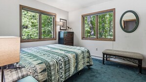 A bedroom with a bed, dresser, bench, two windows with a view of trees, and a round mirror. - A Comfortable & Spacious Retreat in the Powderhorn Village (Ironwood)