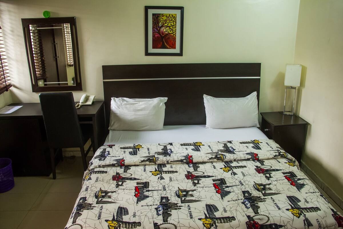 Habitación de hotel con cama, escritorio con teléfono, una silla, espejo y un cuadro en la pared.