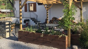 A wooden pergola over a patio area with a stone wall, a grill, and a table with chairs. - Ancienne Fromagerie, a unique chalet, beautifully renovated  in central Samoens  (Samoëns)
