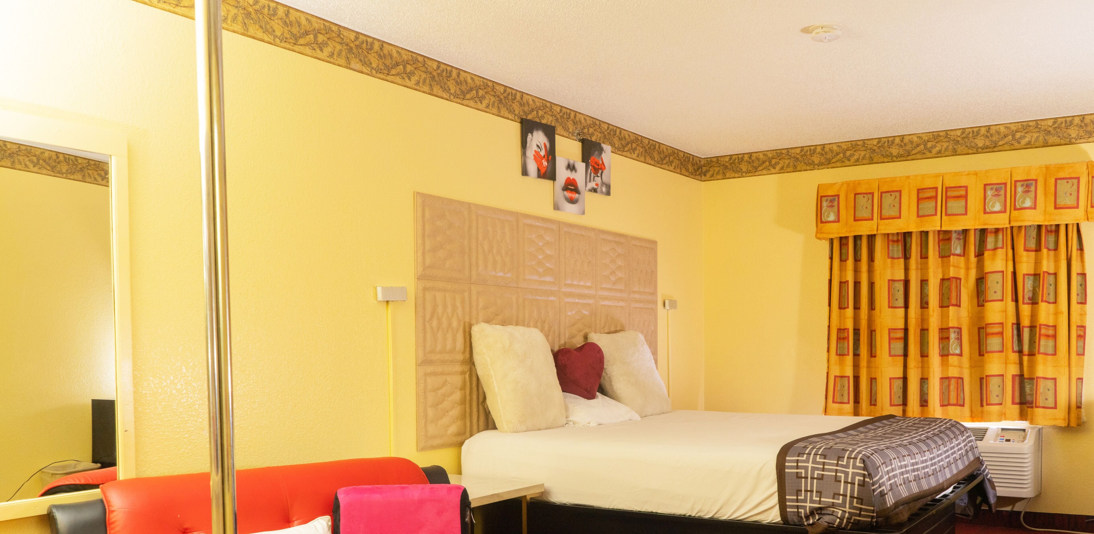 Ein Hotelzimmer mit Bett, einem roten Sessel, einem Schreibtisch mit Computer und einem Fenster mit gelben Vorhängen.