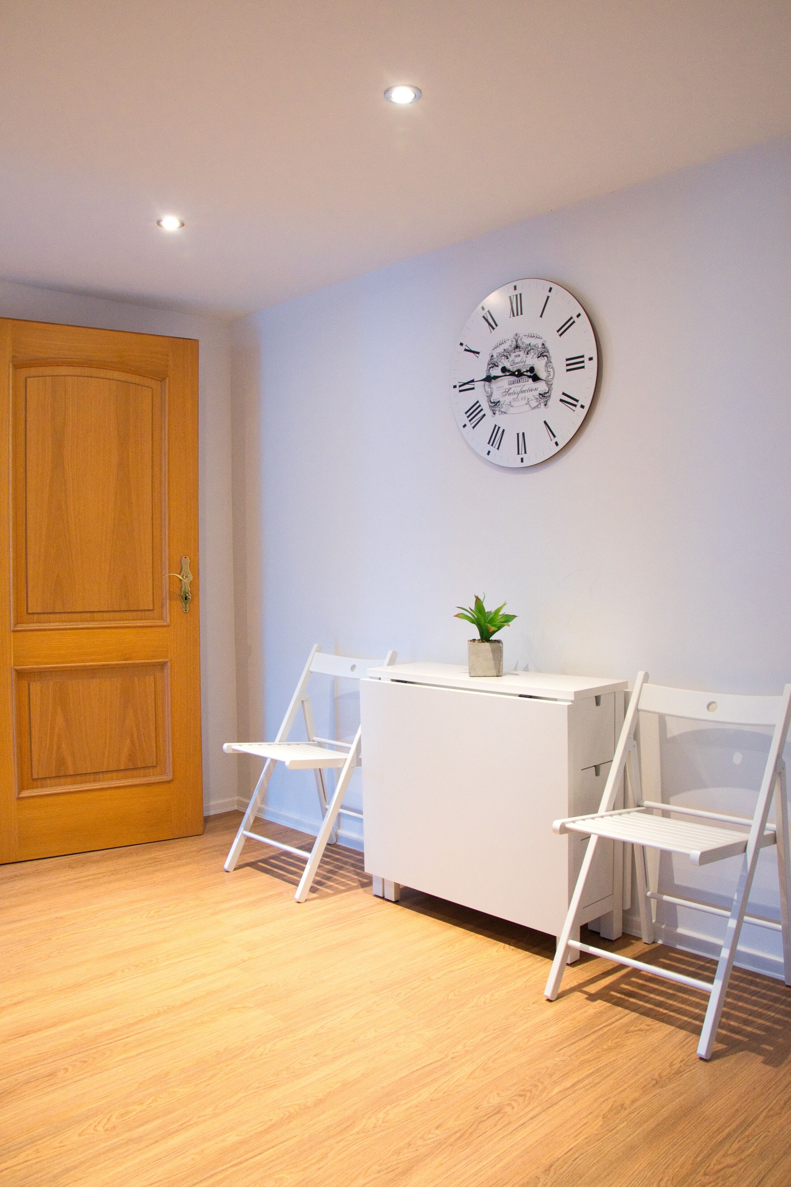 Ein Zimmer mit Holztür, einem weißen Schreibtisch mit einer Uhr und einer kleinen Pflanze.