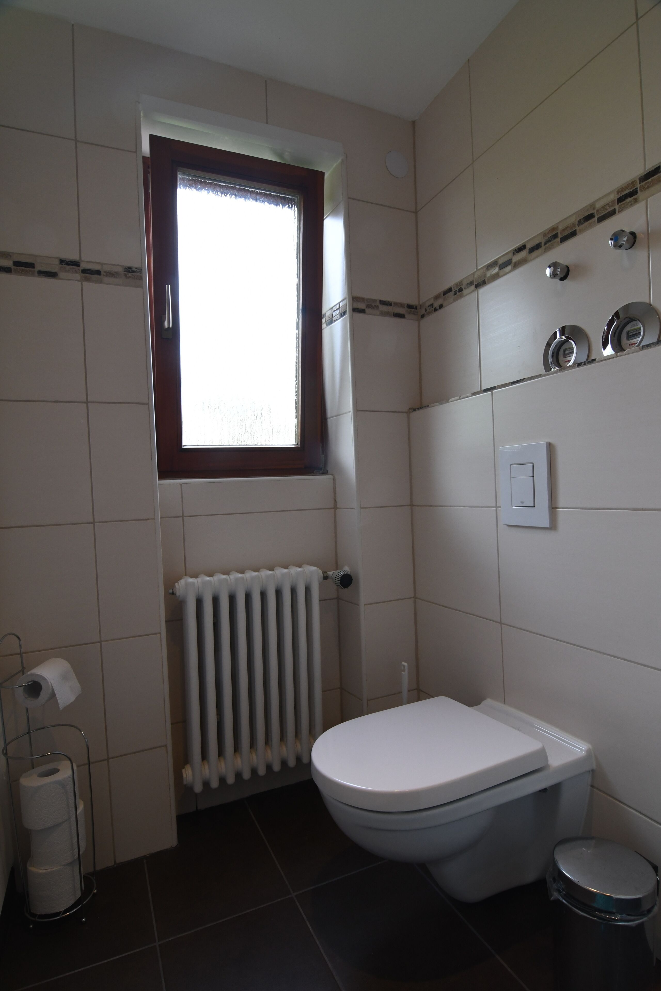 Une salle de bain équipée de toilettes, d’un radiateur, d’une fenêtre et d’une petite poubelle.