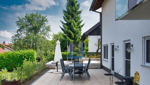 Een terras buiten met een tafel, stoelen, een witte parasol en een brandblusser aan de muur.