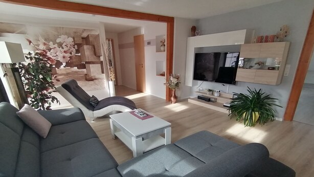 Ein modernes Wohnzimmer mit einer grauen Eckcouch, einem weißen Couchtisch und einem an der Wand befestigten Fernseher. Üchtis sind vorhanden und eine Treppe führt zu einer weiteren Ebene.