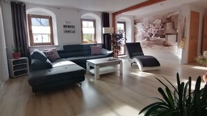 Ein modernes Wohnzimmer mit einem Ecksofa, einem Couchtisch und einem Sessel. Zwei Fenster mit Vorhängen, eine Wandaircondition und eine Wand mit floralem Muster sind ebenfalls zu sehen.