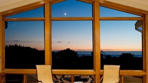 Une pièce dotée de grandes fenêtres offrant une vue sur le coucher de soleil et un croissant de lune. On y aperçoit deux fauteuils en osier au premier plan.