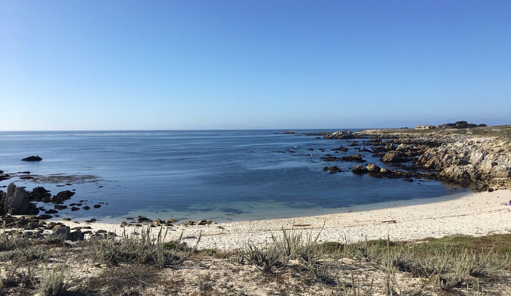 Une plage côtière bordée d’une eau d’un bleu limpide, d’une côte rocheuse et d’une végétation clairsemée.
