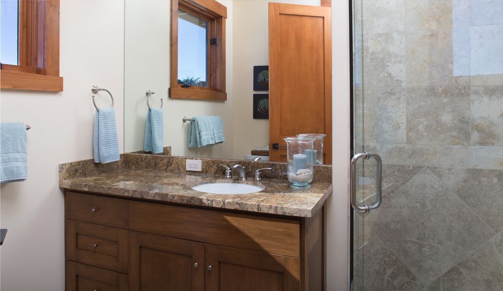Une salle de bain dotée d’un comptoir en granit, d’un lavabo, d’un miroir et d’une cabine de douche en verre.
