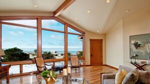 Il s’agit d’une pièce spacieuse, dotée de grandes fenêtres qui offrent une vue sur la plage et l’océan. On y trouve un plafond en bois, une table basse en verre et un coin salon confortable avec un canapé et des fauteuils.