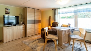 A modern living room with a flat-screen TV, wooden cabinets, a dining table with chairs, and a large window with curtains. - "Kleine Küstenpause" - Apartment in the outbuilding (Wlan) (Garz/Rügen)