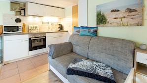 A compact living space with a kitchenette, a sofa with decorative pillows, and a beach-themed painting on the wall. - "Kleine Küstenpause" - Apartment in the outbuilding (Wlan) (Garz/Rügen)