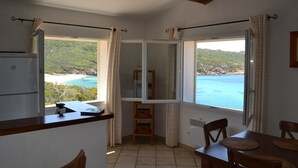 A room with a kitchen area, a dining table with chairs, and large windows offering a view of a beach and sea.