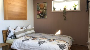 Een slaapkamer met een bed, een houten hoofdbord, een raam met planten en een kunstwerk aan de muur.